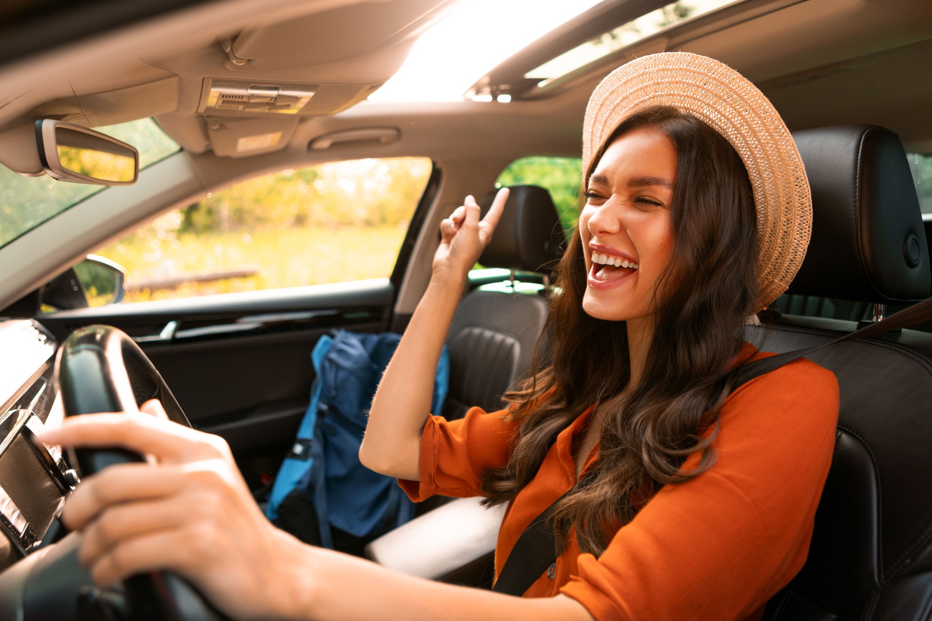 Une touriste ravie profitant d’un trajet en voiture seule, écoutant de la musique, chantant des chansons et dansant tout en conduisant une voiture avec un sac à dos à proximité sur le siège passager